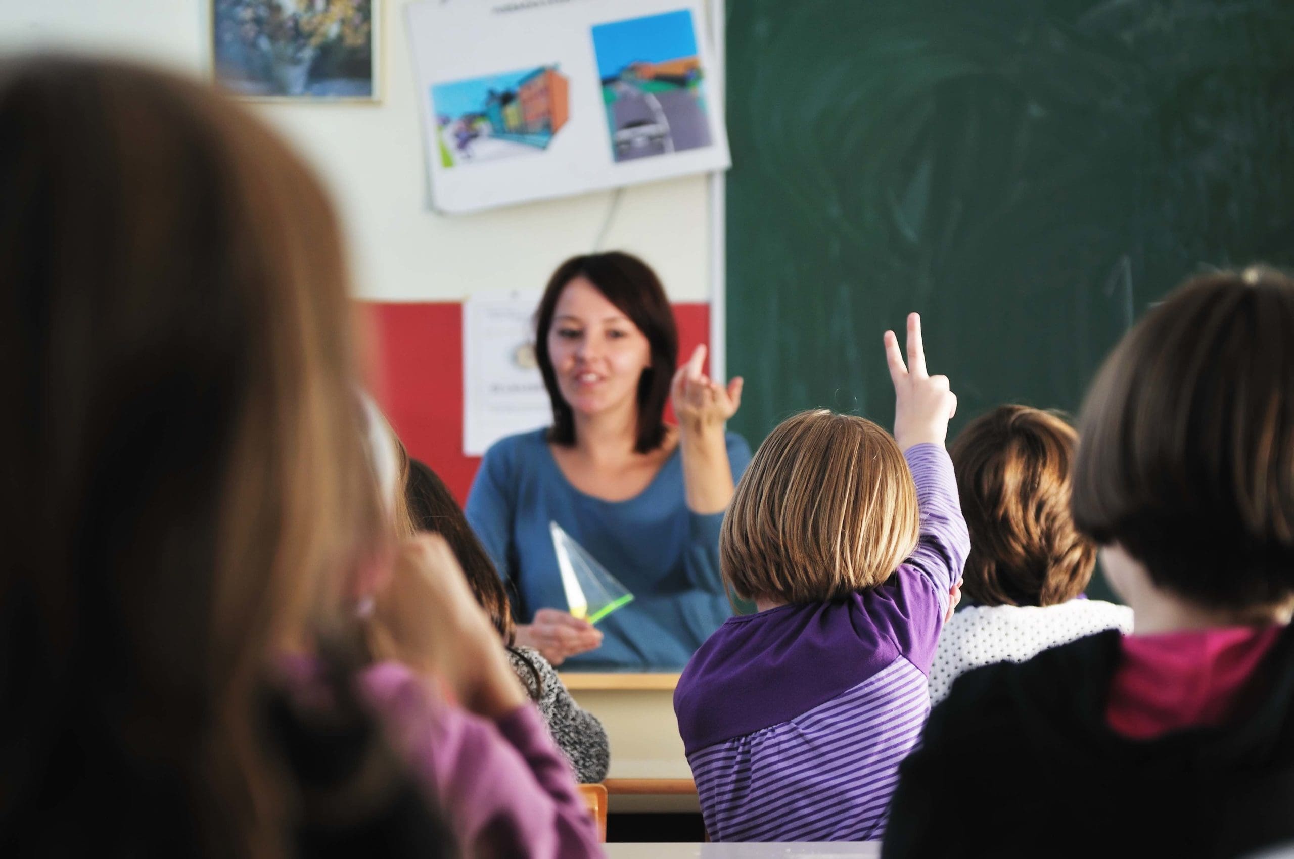Elementary school teacher sitting at her desk calling on a young student who has her hand raised.