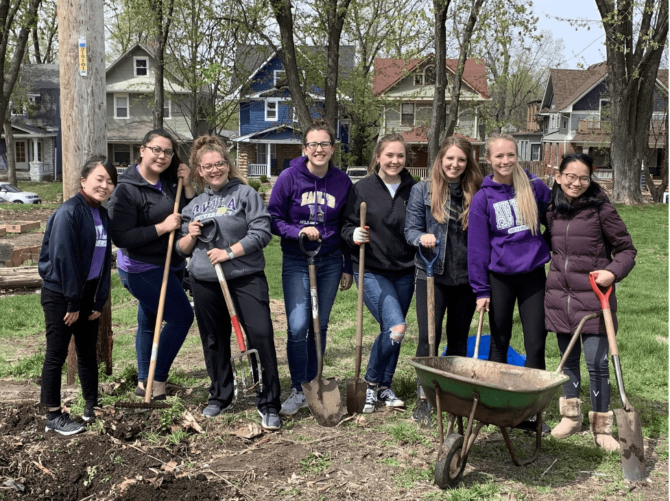 Eight students with shovels, rakes, forks, and wheelbarrow gardening at St. James